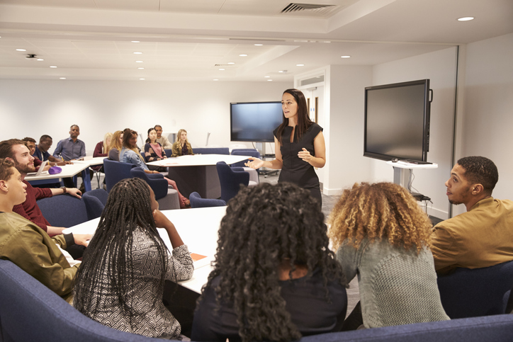 A female teacher stands in front of a classroom of university students and speaks to them